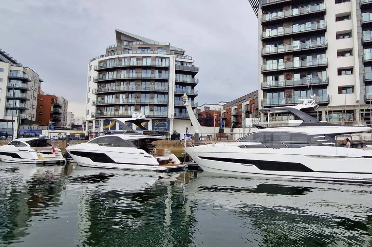 Yachts docked in a marina with modern apartment buildings in the background under a cloudy sky.