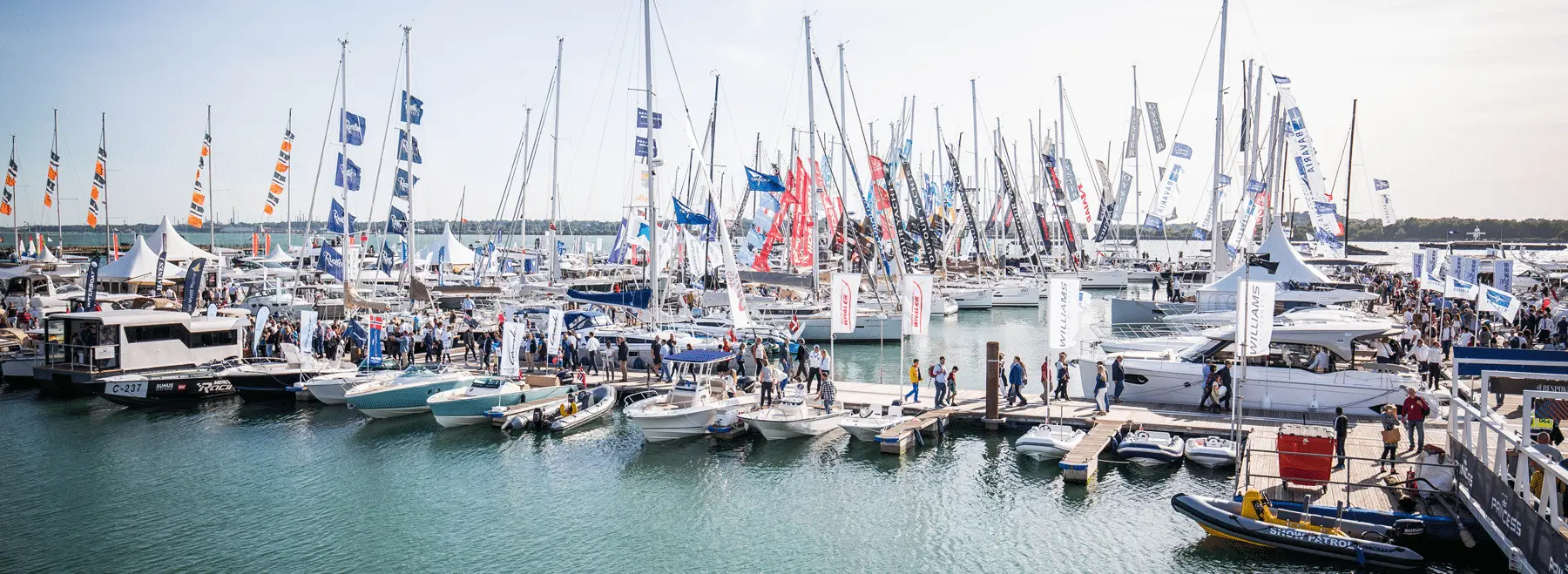Crowded marina during a boat show, featuring numerous boats, flags, and people exploring the exhibits and waterfront.