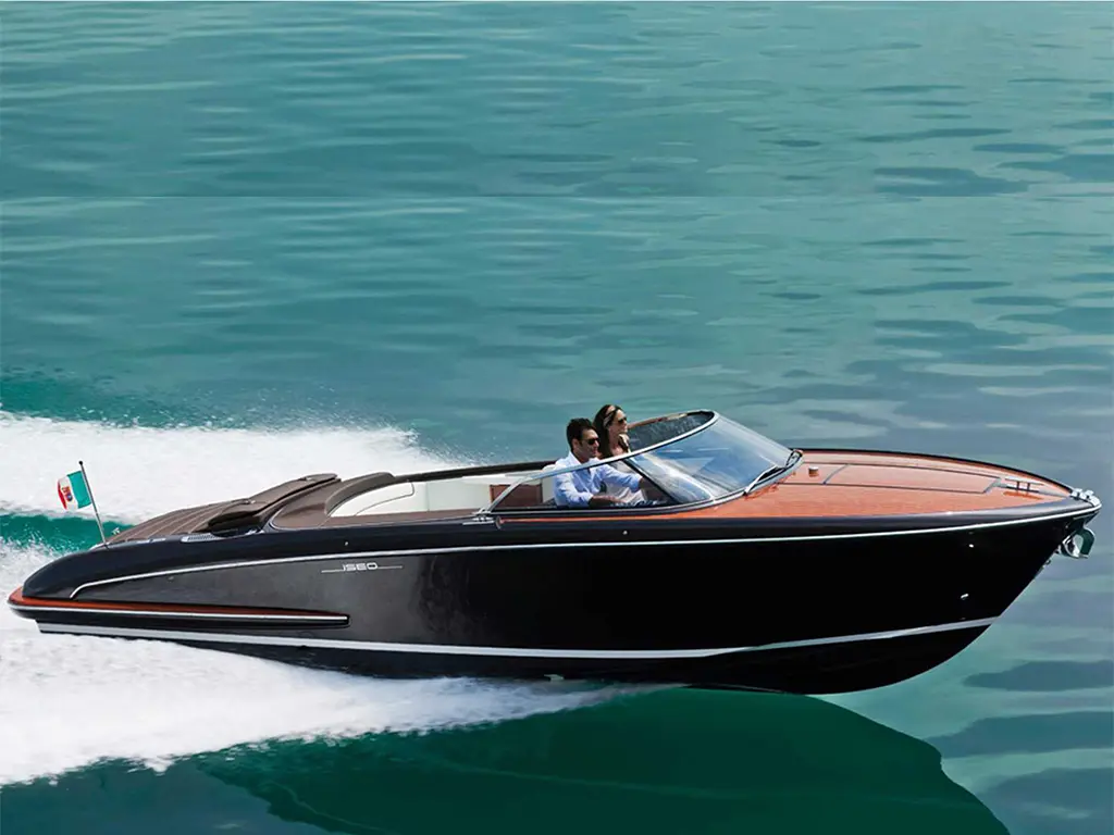 A stylish black speedboat with wooden accents speeds across turquoise waters, with two passengers enjoying the ride.