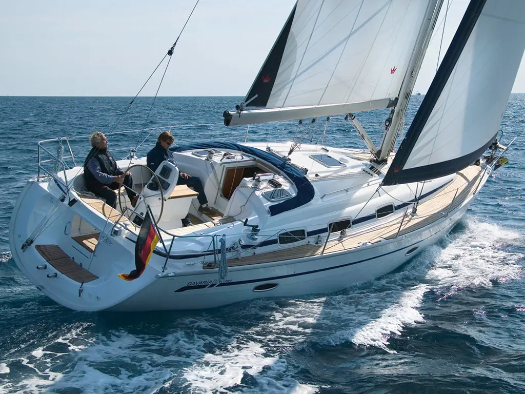A Bavaria sailing yacht glides through turquoise waters, with two men aboard adjusting sails under a clear sky.