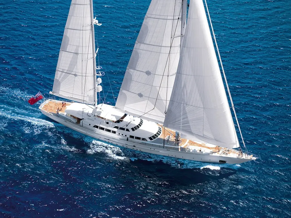 Aerial view of a large sailing yacht gliding through deep blue waters, with billowing white sails and a red ensign flag.