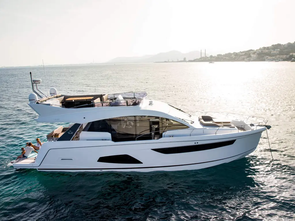 A sleek white yacht cruising on calm waters, with people relaxing on the back, set against a sunny coastline.