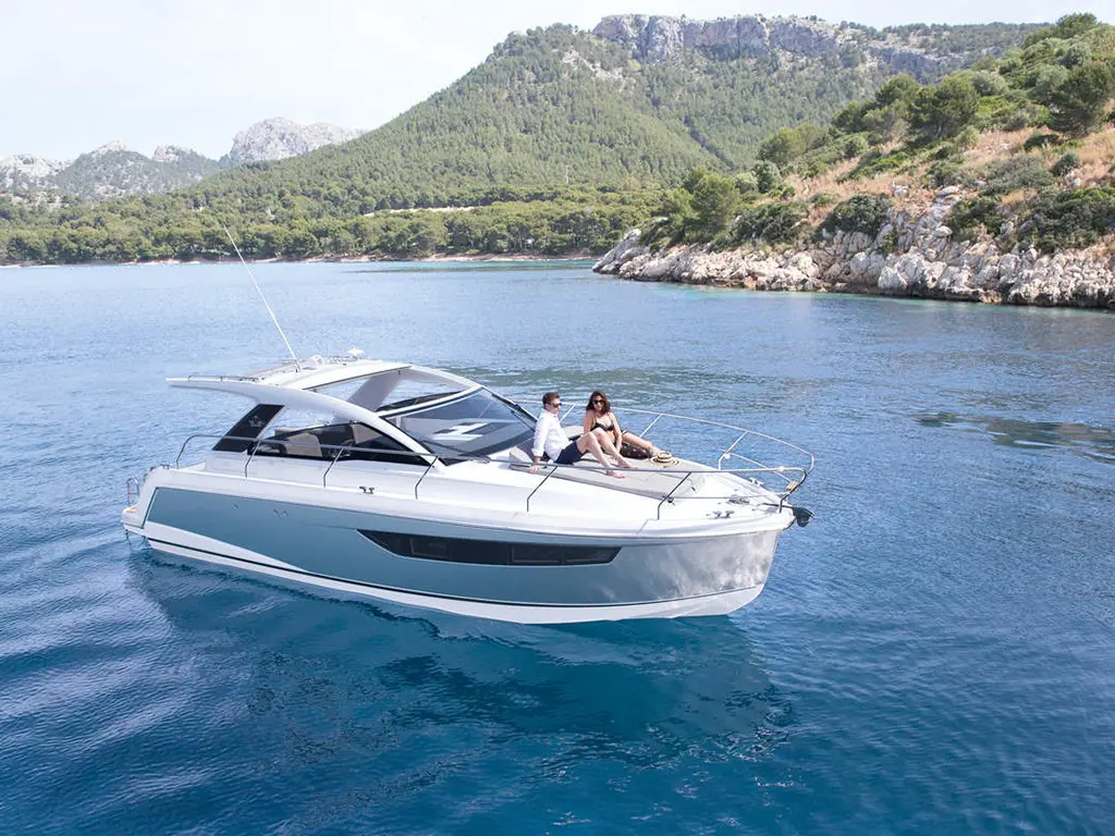 A sleek motorboat floats on clear blue water, with two people relaxing on the deck and mountainous greenery in the background.