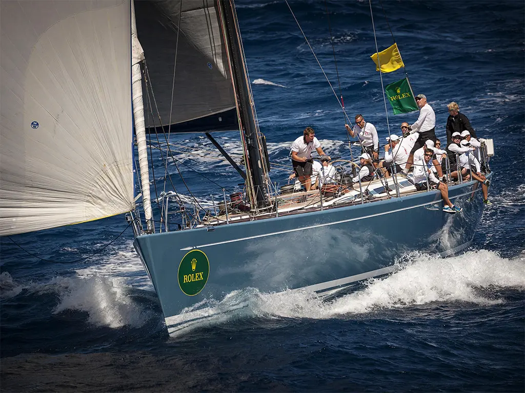 A sleek sailing yacht with a green Rolex logo, navigating choppy seas, with a crew in white shirts actively managing the sails.