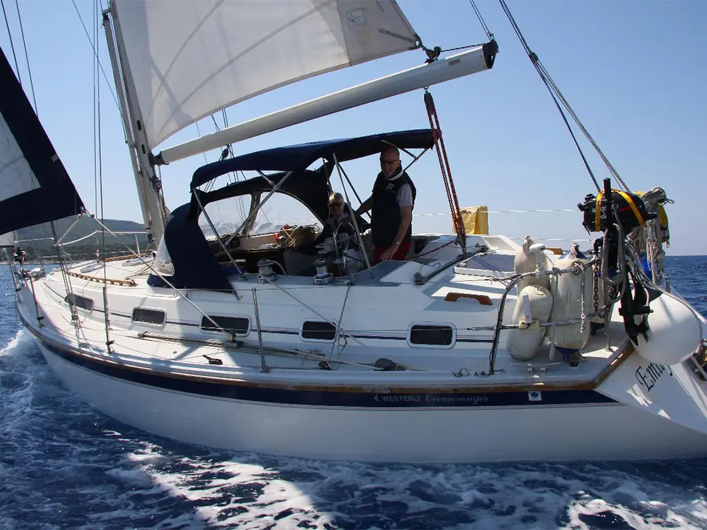 Sailing yacht with two people on board, one steering and the other seated, surrounded by clear blue water and visible sails.