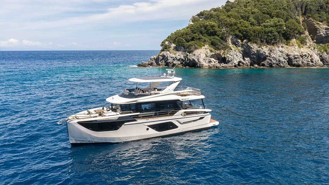 Luxury motor yacht cruising beside a rocky, tree-covered Mediterranean coastline in clear blue water.