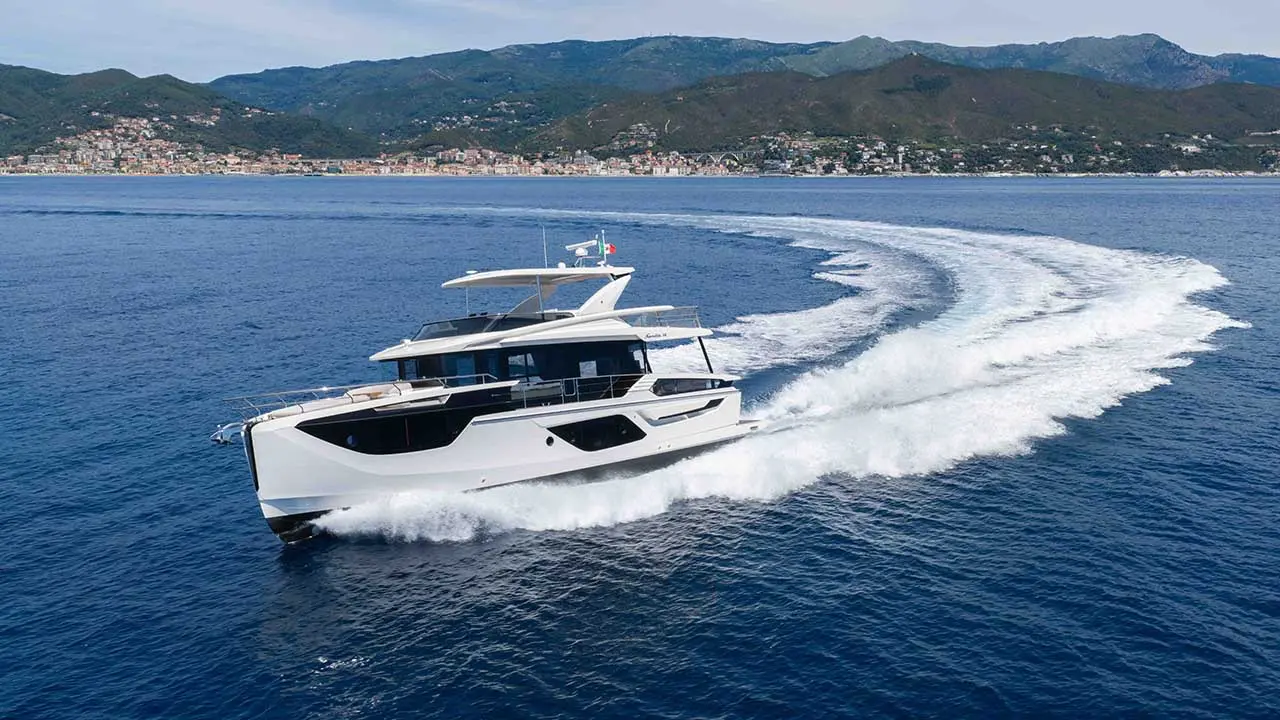 White motor yacht speeding across blue sea, leaving a wake, with a coastal town and mountains behind.