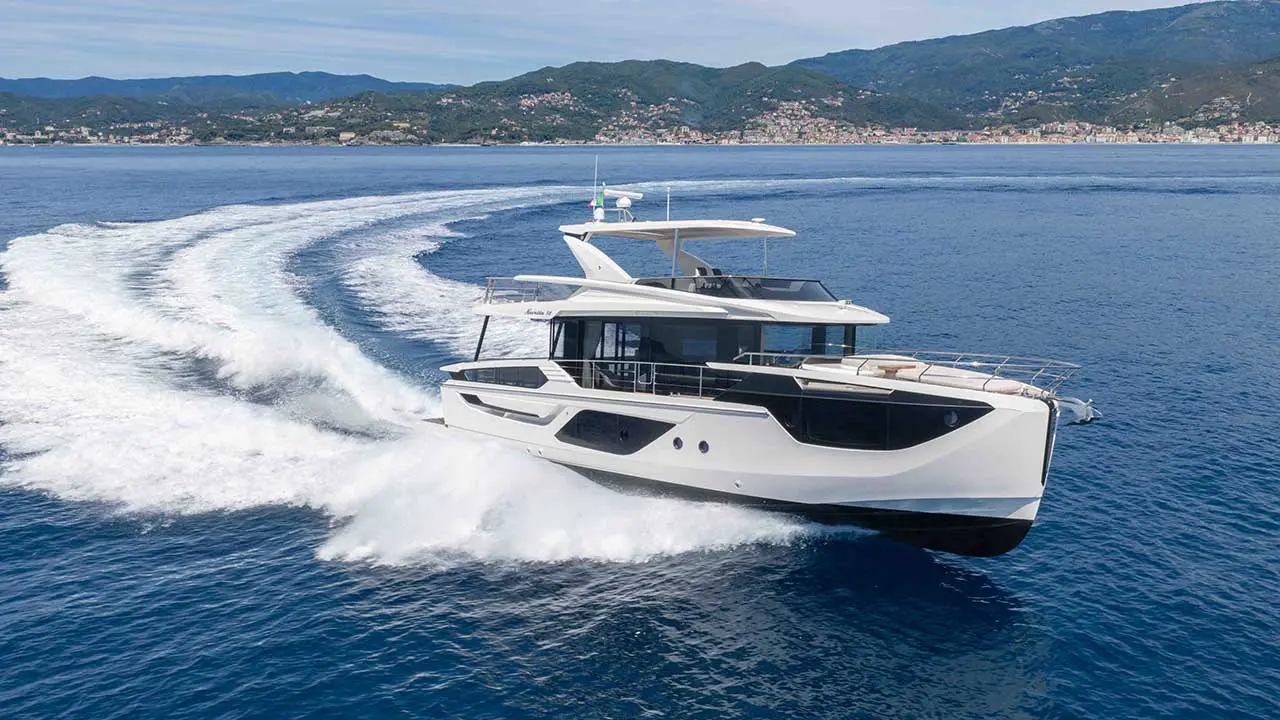 White motor yacht speeding across blue sea, leaving a wake, with a mountainous coastal town in the background.