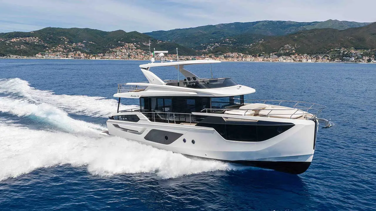 Luxury motor yacht cruising through blue water, with a coastal town and forested mountains in the background
