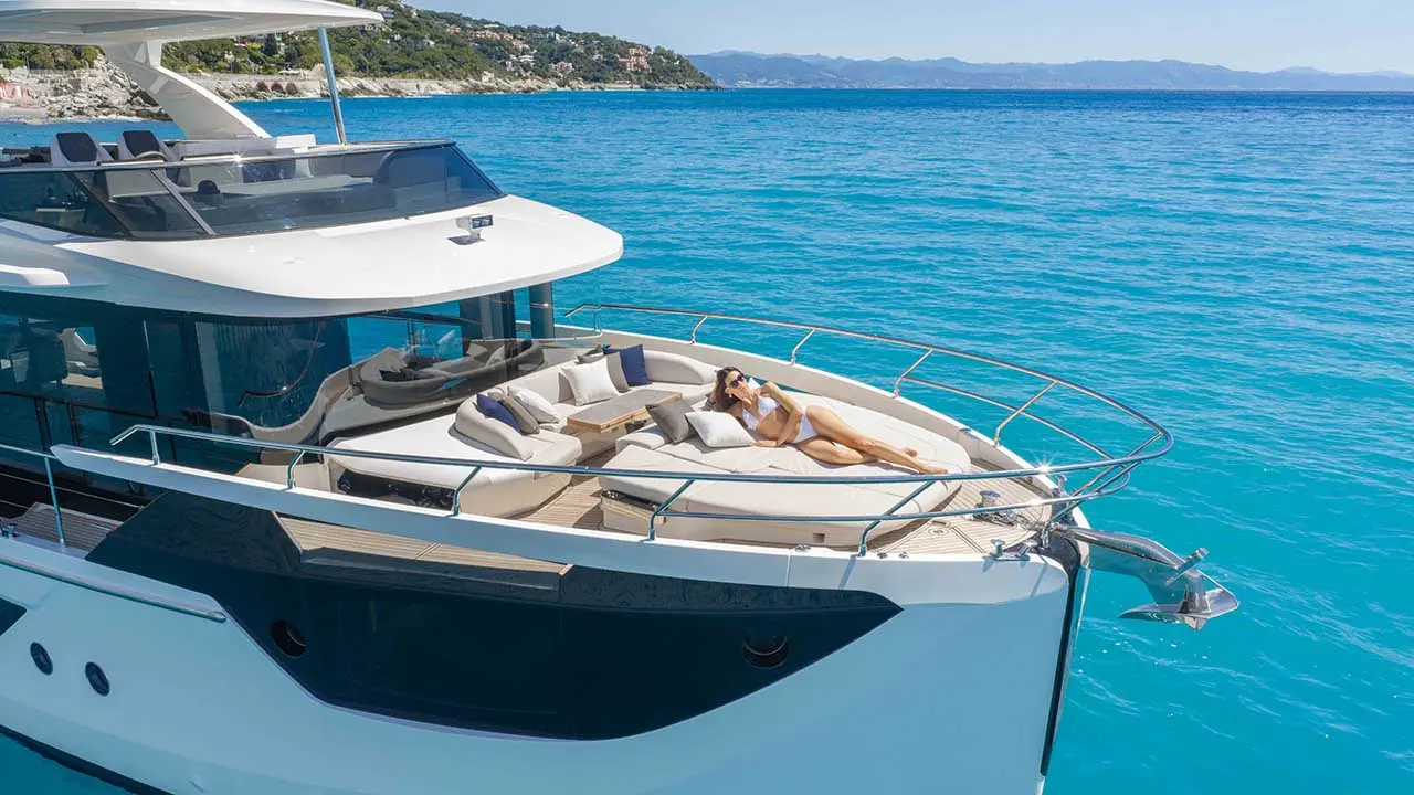 Luxury yacht with a woman sunbathing on deck in turquoise Mediterranean waters near a rocky coastline.
