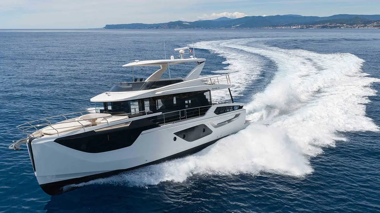 White luxury motor yacht cruising across the sea, leaving a foamy wake, with a mountainous coastline in the background.