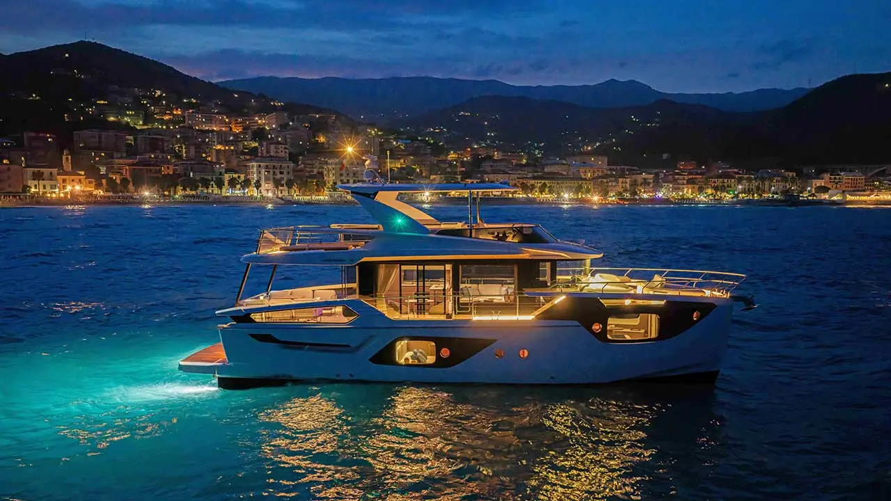 Luxury motor yacht cruising at dusk, with a brightly lit lakeside town and mountains in the background.