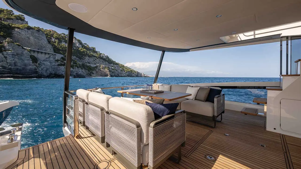 Luxury yacht aft deck with cream lounge seating, dining table and ocean views of rugged coastal cliffs.