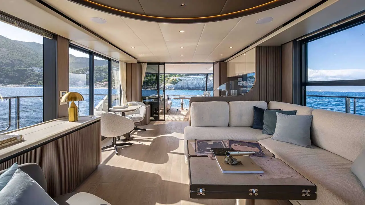Luxury yacht interior with lounge, dining area and panoramic windows overlooking blue sea and coastal mountains.