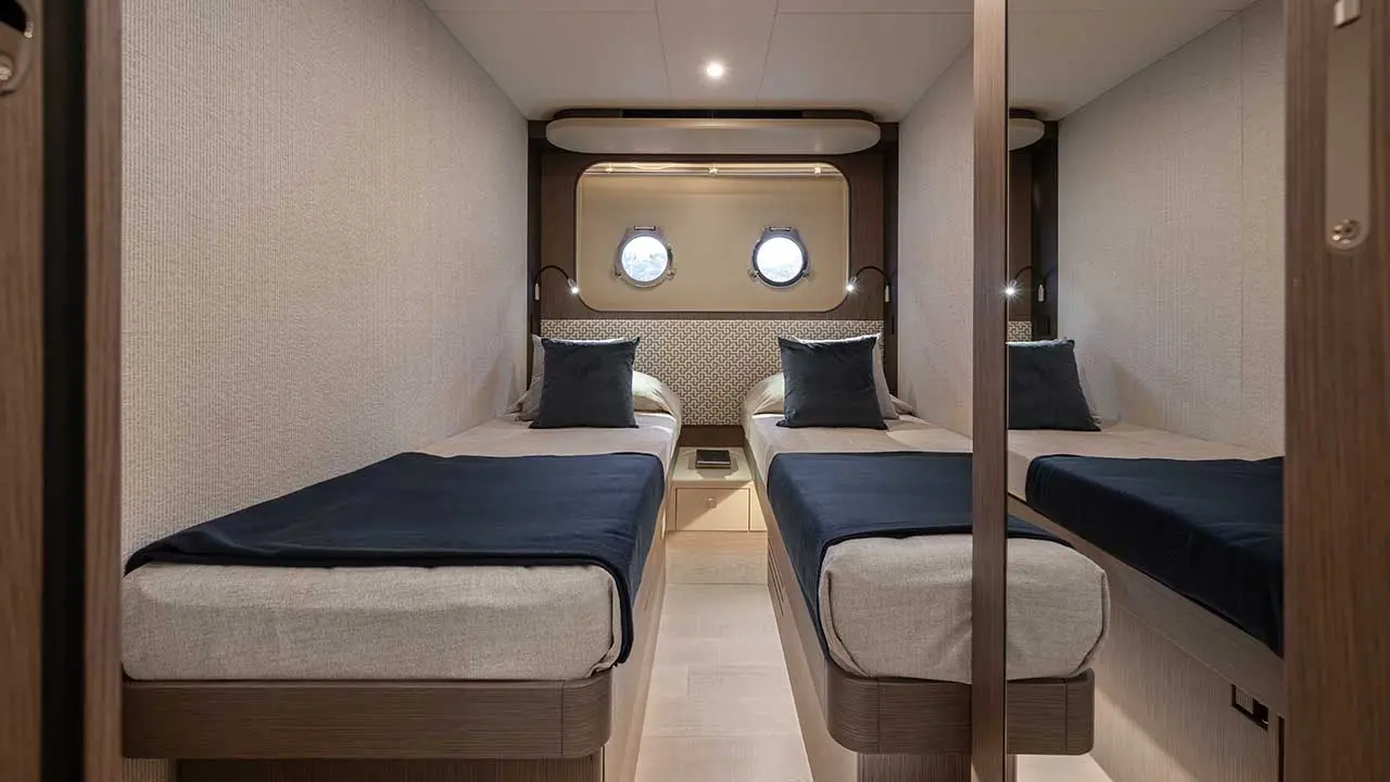 Modern twin-bed yacht cabin with porthole windows, navy blankets, bedside table and mirrored wardrobe.