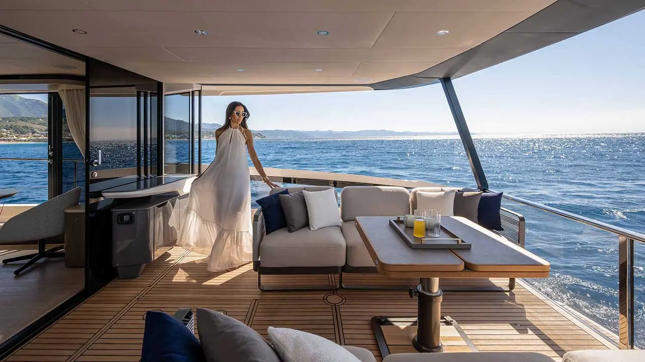 Woman in a white dress on a luxury yacht deck overlooking the sparkling sea and mountainous coastline.