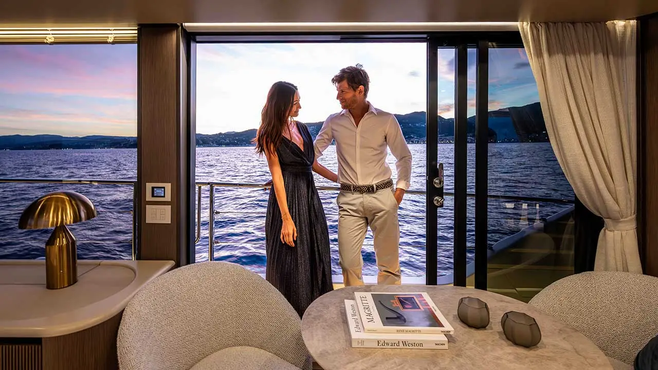 Couple posing in a luxury yacht saloon overlooking a lake and mountains; books titled “Magritte” and “Edward Weston” on table.
