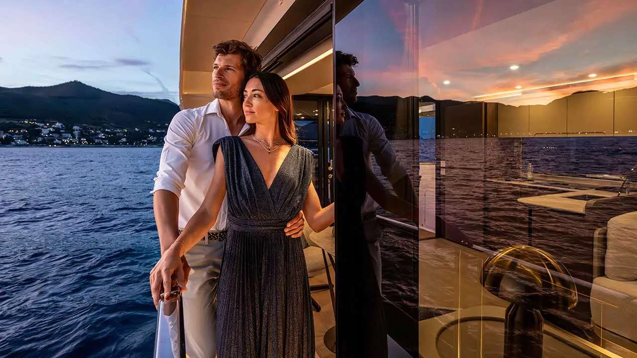 Couple on a luxury yacht overlooking a lake and mountains at sunset, with the stylish interior reflected in glass panels.
