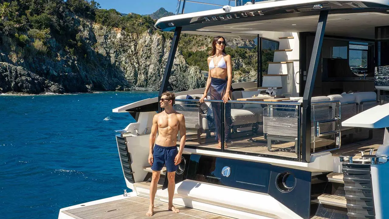 Couple on the deck of luxury yacht “ABSOLUTE” in a sunny blue-water cove surrounded by rocky cliffs.