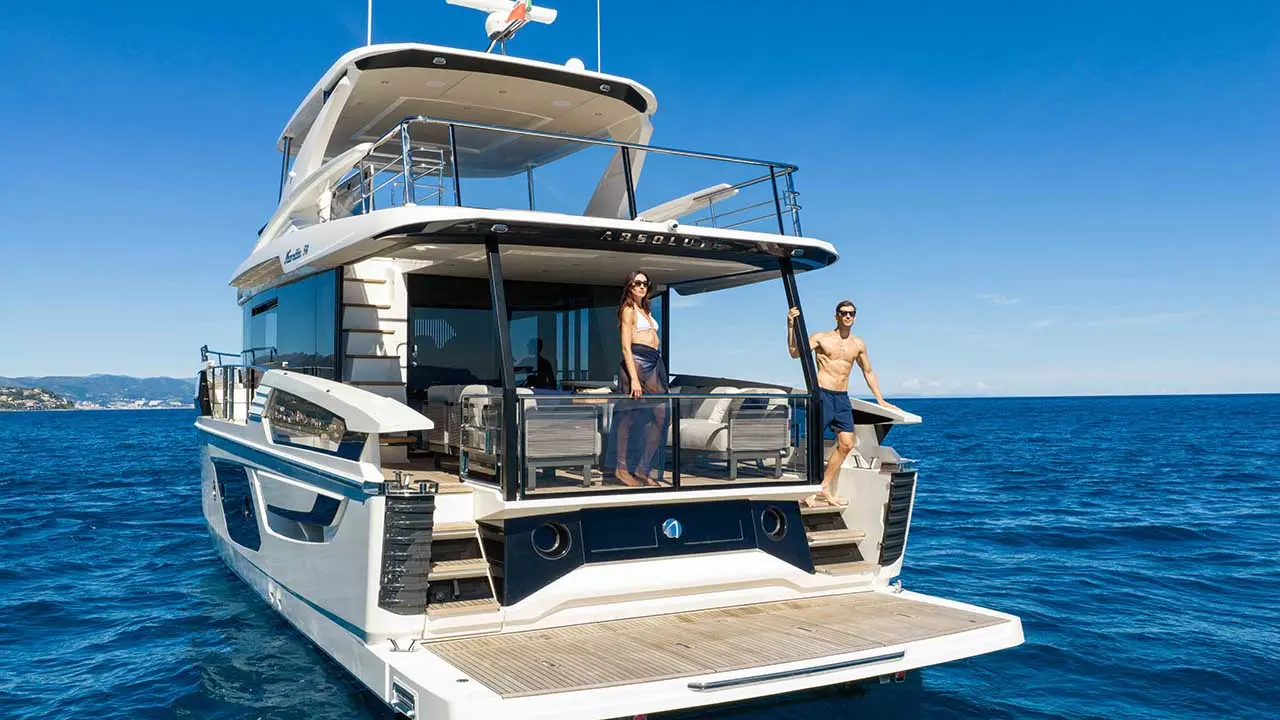 Luxury motor yacht named “ABSOLUTE” with two people aboard on blue sea near a coastal landscape.