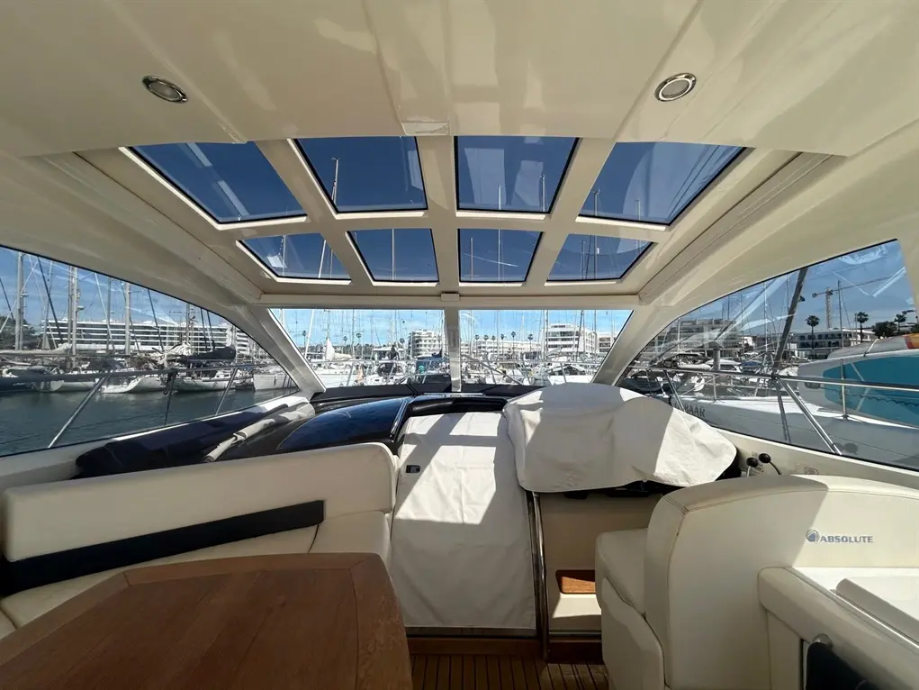 Interior view of a yacht featuring large windows, a wooden table, and the marina in the background with parked boats.