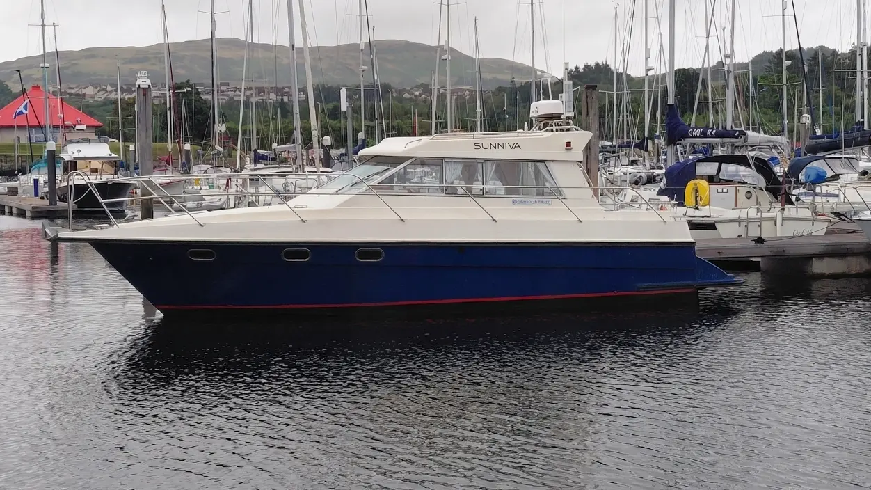 Motor yacht named “SUNNIVA” moored in a marina, surrounded by sailboats, with green hills in the background.