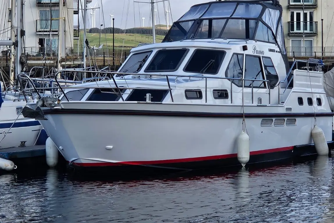 White motorboat named 'Puma' docked at a marina, surrounded by other boats and a tranquil water reflection.