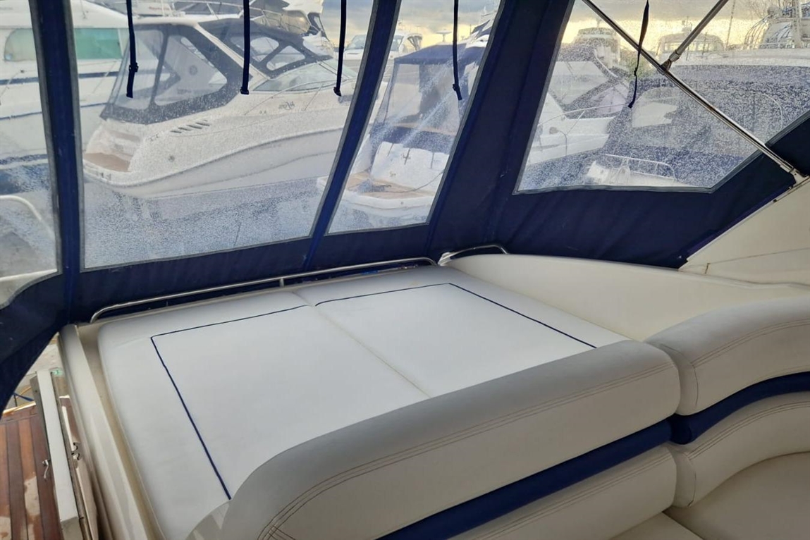 Interior of a boat showing a white cushioned seating area with blue trim, framed by clear vinyl windows and parked boats outside.