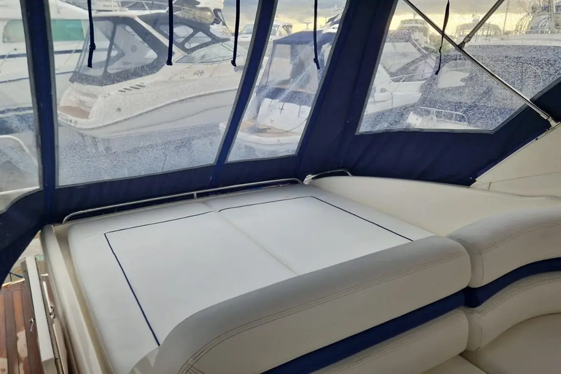 Interior of a boat showing a white cushioned seating area with blue trim, framed by clear vinyl windows and parked boats outside.