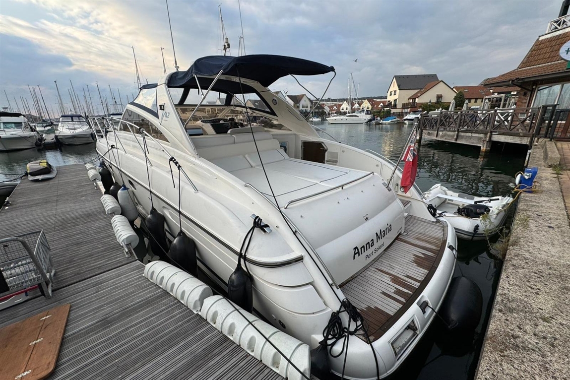 A white boat named "Anna Maria" docked at a marina, with nearby boats and a cloudy sky reflecting in the water.