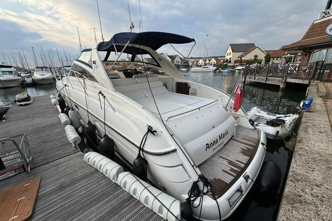 A white boat named "Anna Maria" docked at a marina, with nearby boats and a cloudy sky reflecting in the water.