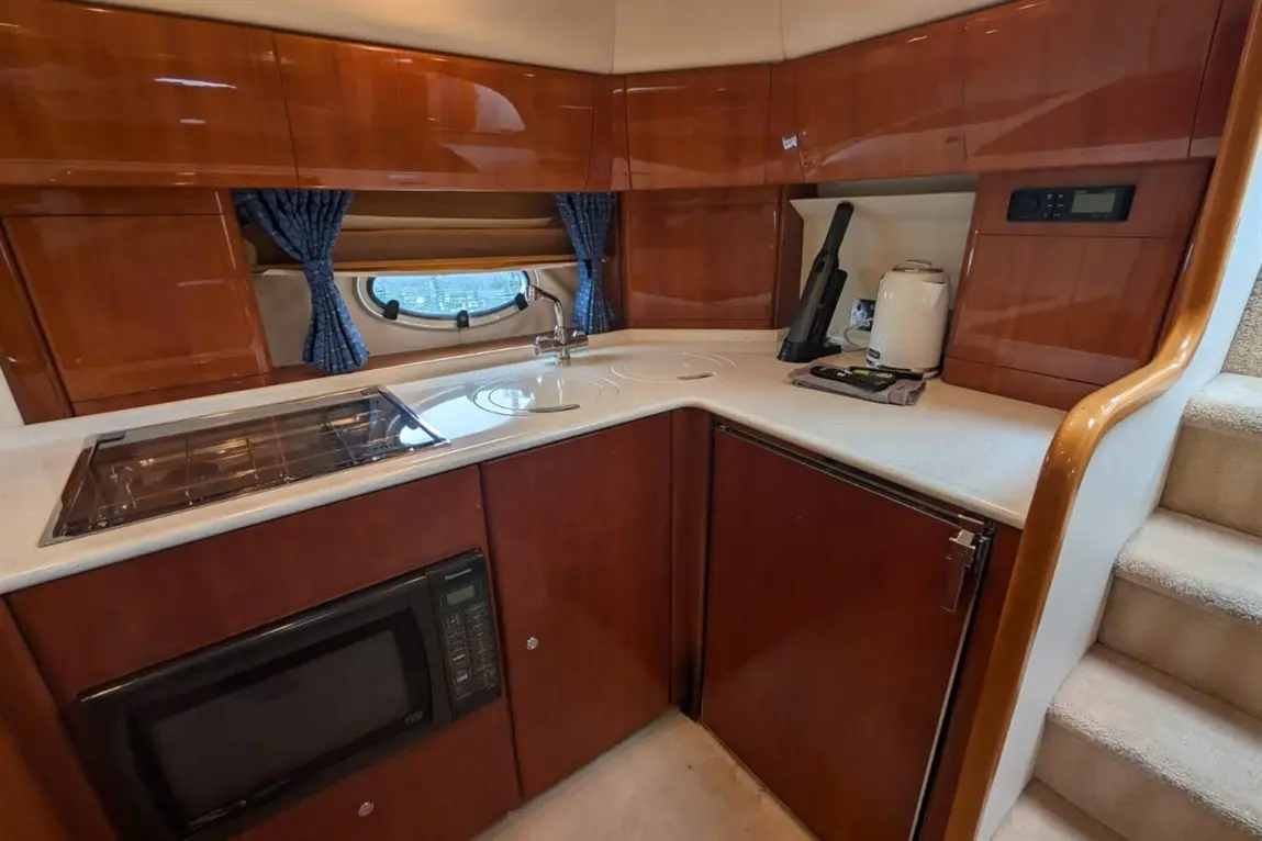 Modern yacht kitchen with wood cabinetry, a microwave, stove, sink, kettle, and blue curtains near a round window.
