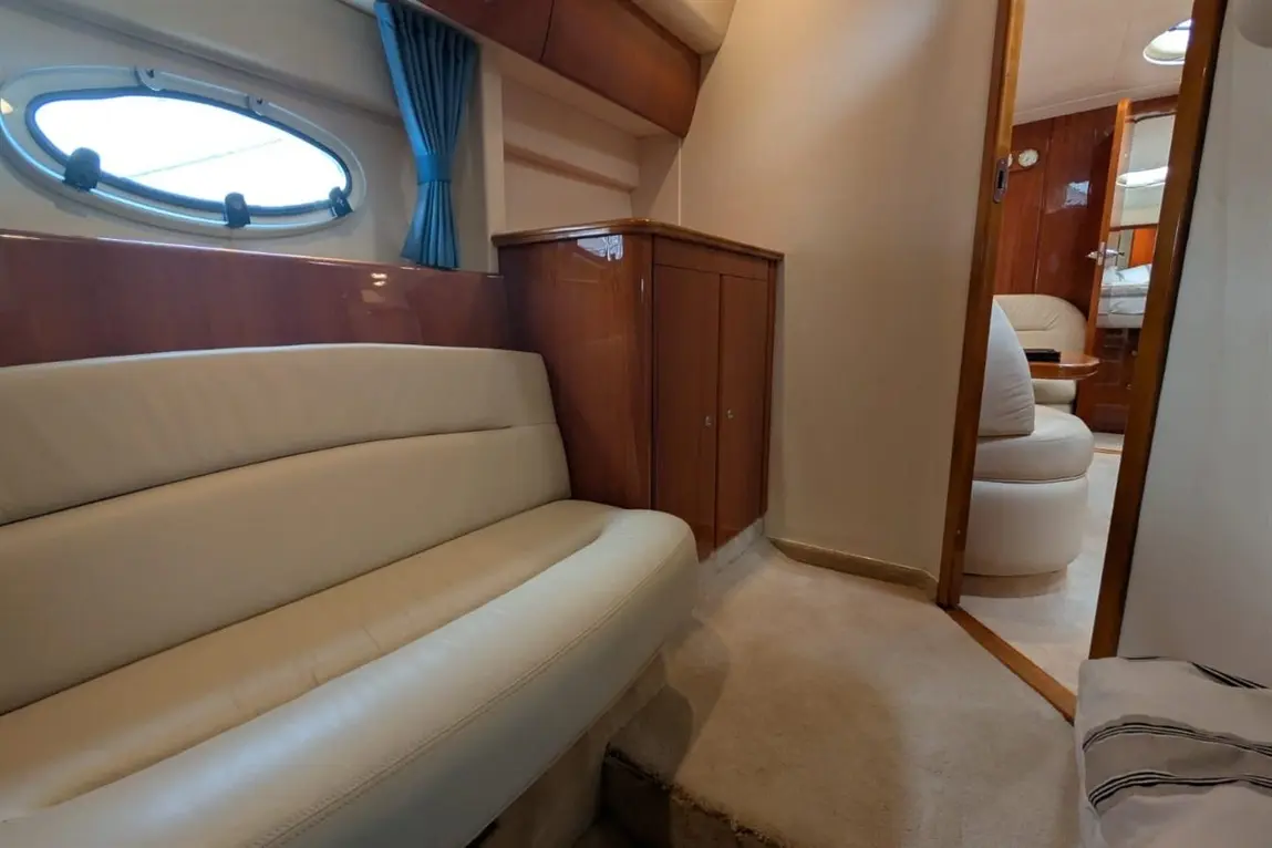Interior of a yacht featuring a white leather sofa, wooden cabinetry, and an oval window with blue curtains.
