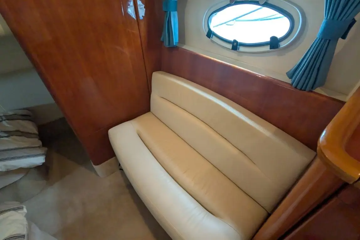 Interior view of a boat cabin featuring a light leather sofa and wooden wall, with a round window and blue curtains.