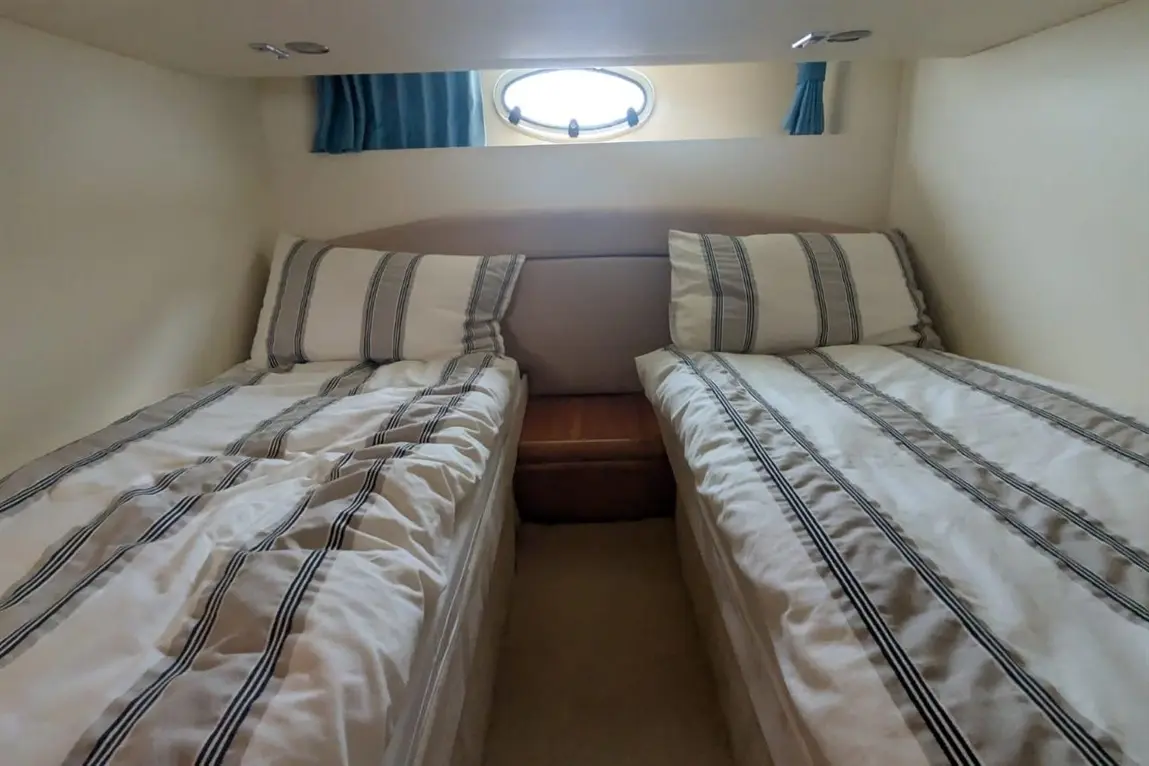 Two single beds with striped bedding, a wooden bedside table, and a porthole view in a compact cabin space.