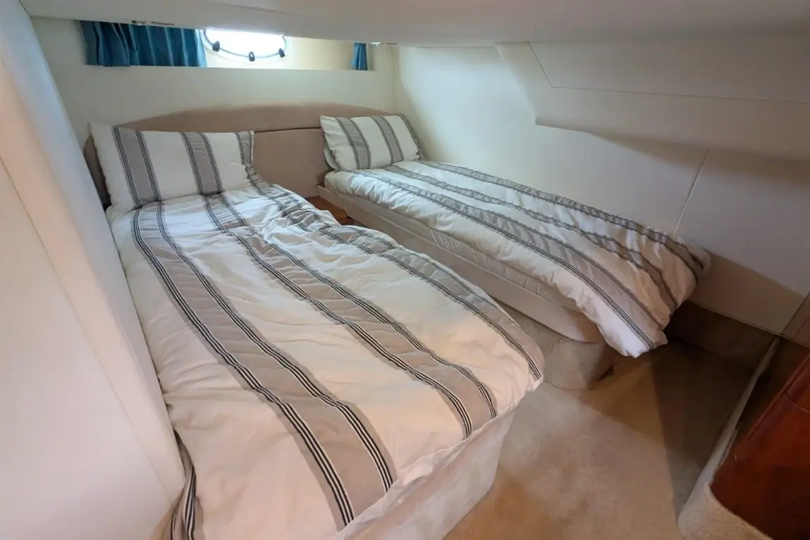 Two single beds with striped bedding in a narrow cabin, featuring a porthole window and beige wall panels.