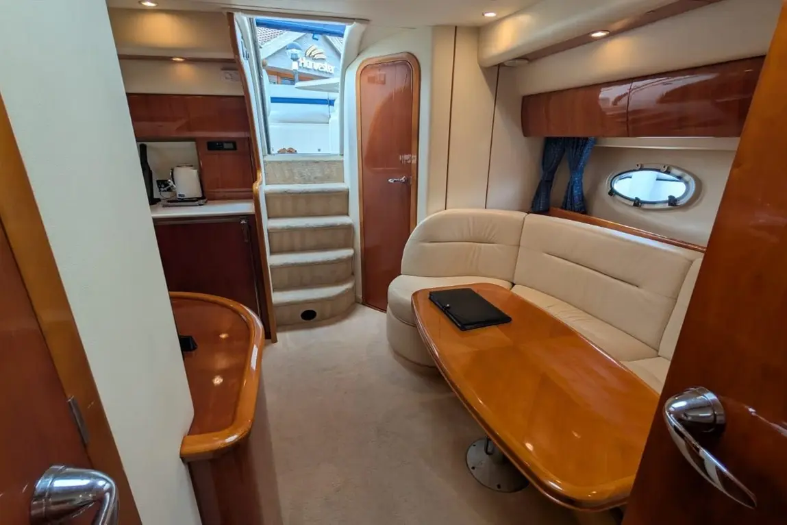 Interior of a boat featuring a seating area, wooden table, kitchenette, and steps leading above, with a window showing outdoor views.