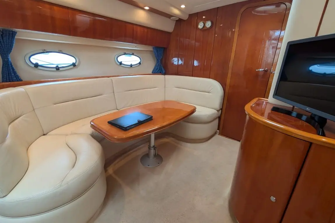 Luxurious boat interior featuring a leather sofa, round wooden table, television, and porthole windows with blue curtains.