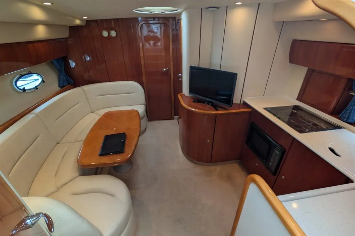 Interior of a boat featuring a cream-coloured sofa, wooden dining table, television, and compact kitchen area.