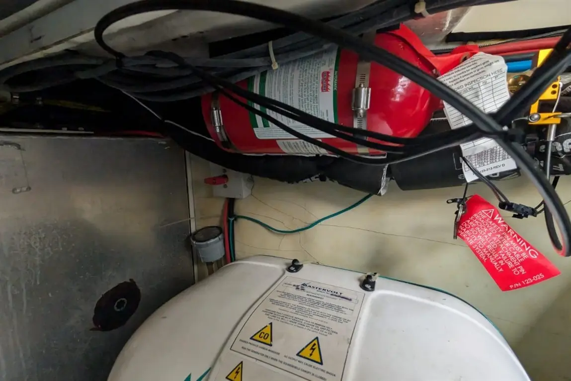 Red fire extinguisher mounted above a white Mastervolt unit with warning labels and various electrical wires in a storage compartment.