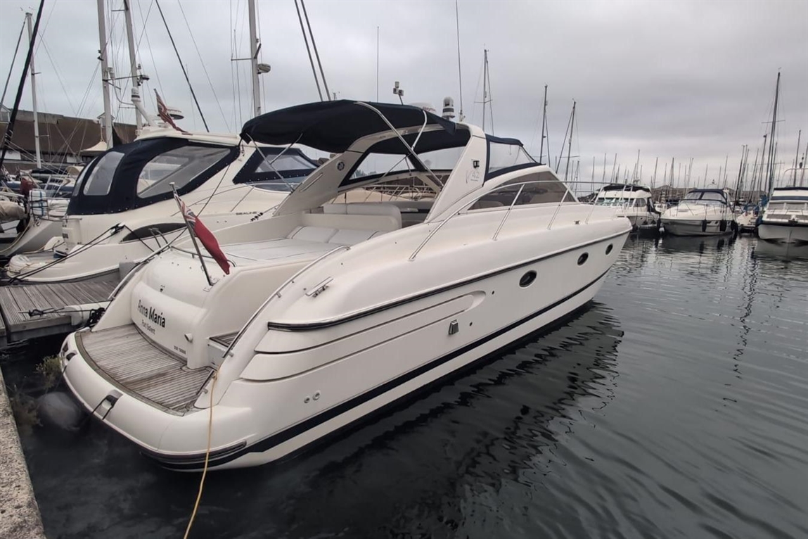 A white motorboat named "Prima Maria" is docked in a marina, surrounded by several other boats under a cloudy sky.