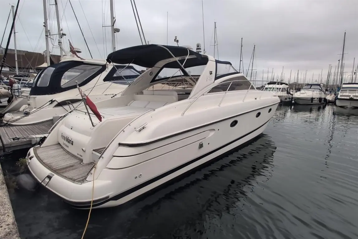 A white motorboat named "Prima Maria" is docked in a marina, surrounded by several other boats under a cloudy sky.