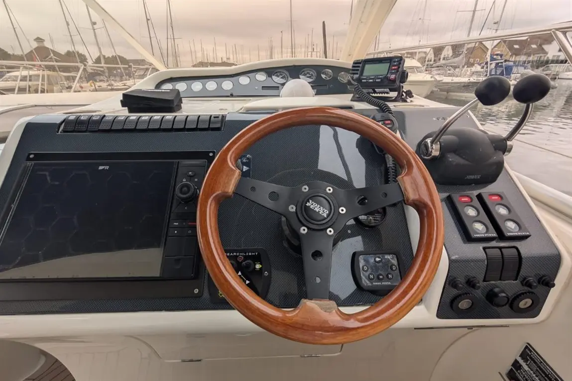 Boat dashboard featuring a wooden steering wheel, navigation display, controls, and surrounding marina scenery.