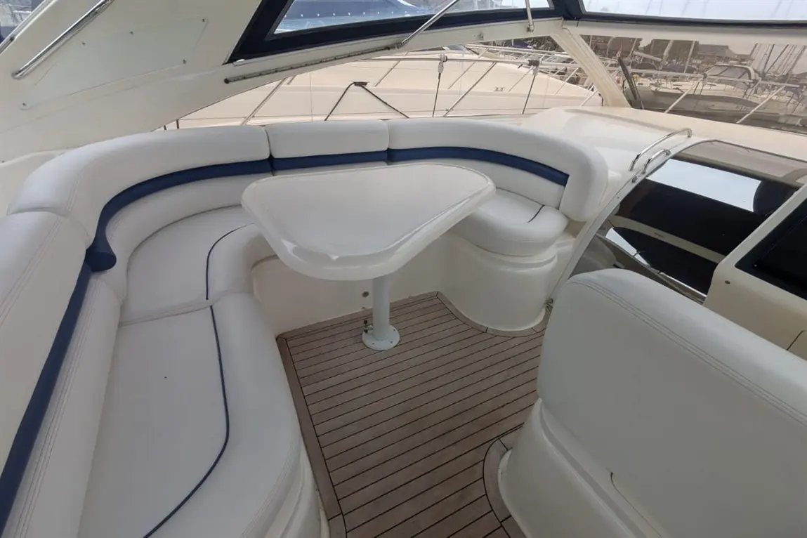 Luxury boat interior featuring a curved white leather seating area and a small table on a wooden floor, with marina views.