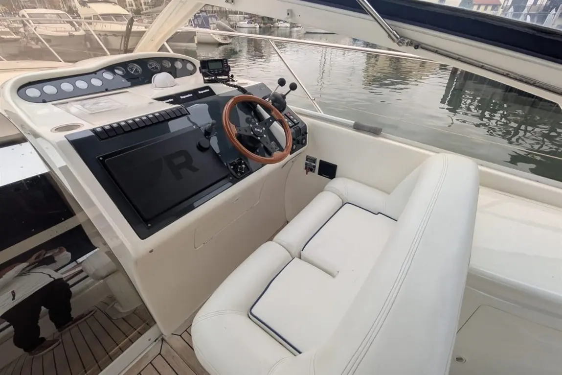 Boat cockpit featuring a wooden steering wheel, control panel, and plush white seating, overlooking a marina.