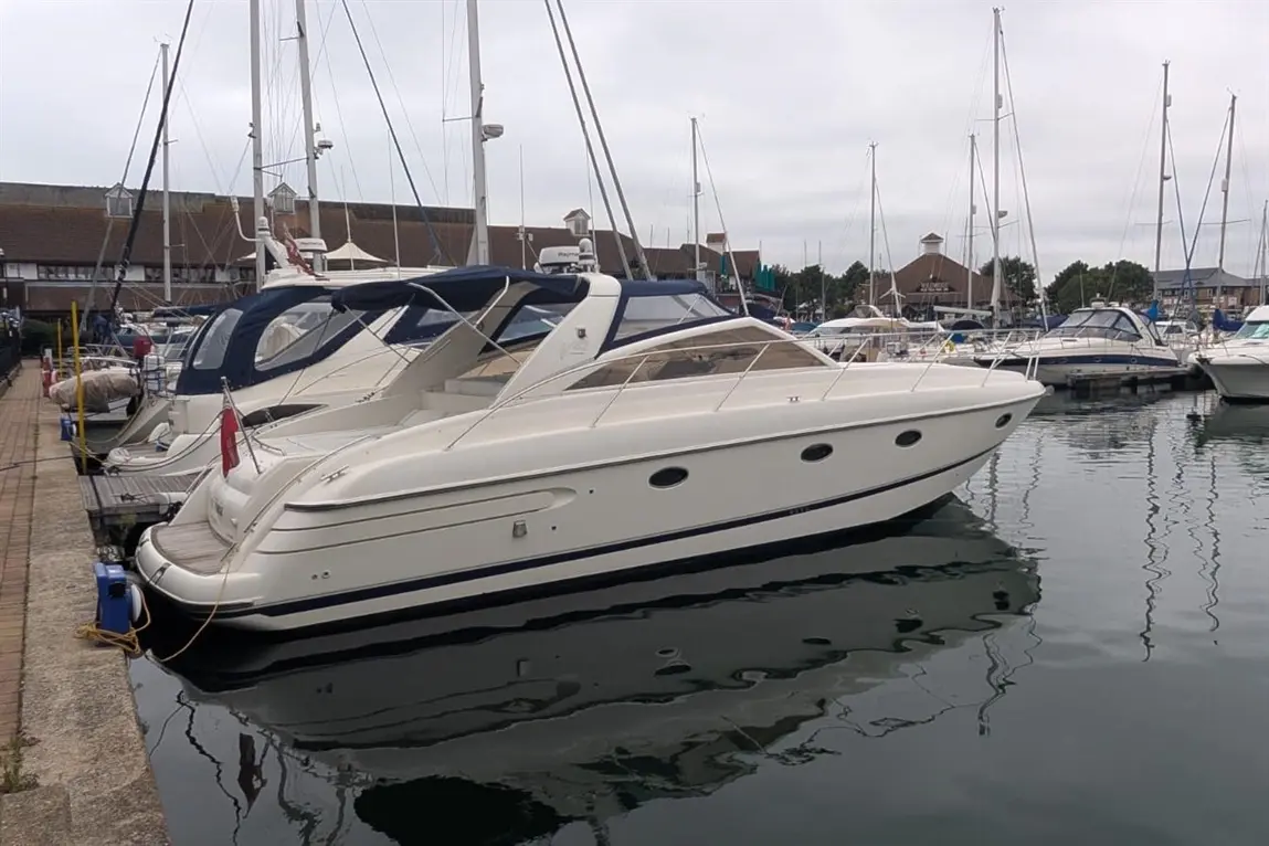 White motorboat docked at a marina beside other boats, with a calm water reflection under a cloudy sky.