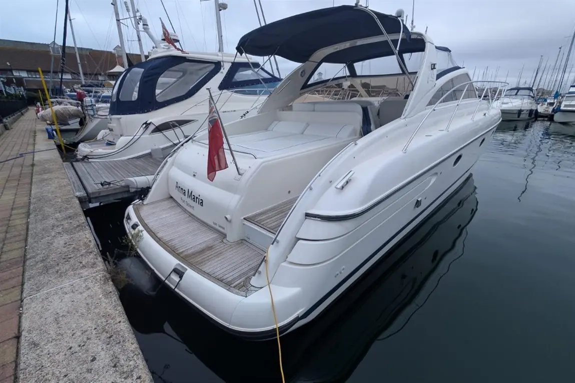 A white motorboat named "Anna Maria" docked at a marina, surrounded by other boats and calm water under a cloudy sky.