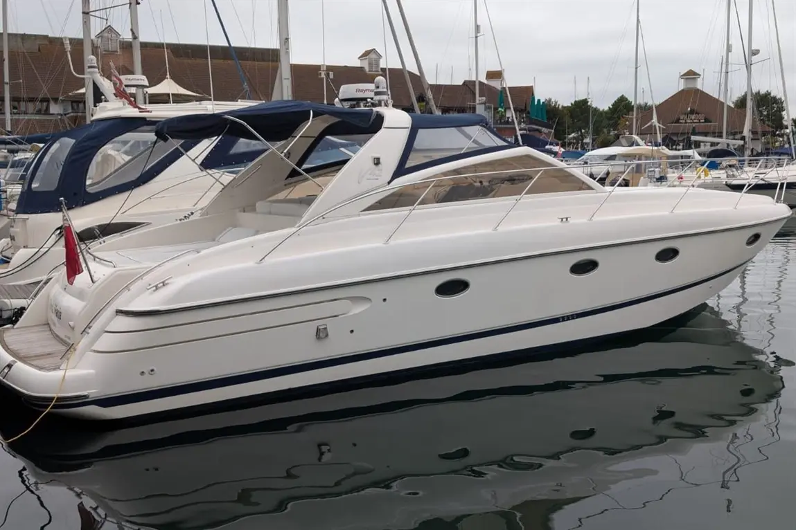 A sleek white motorboat docked at a marina, reflecting in calm water, surrounded by other boats and mooring posts.
