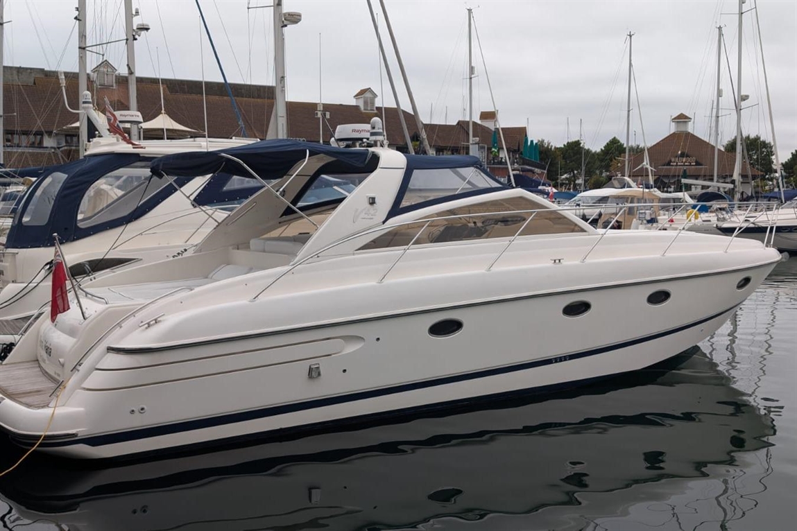 A sleek white motorboat docked at a marina, surrounded by other boats and masts under a cloudy sky.