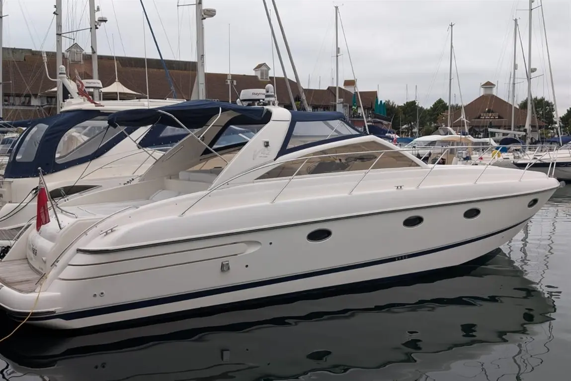 A sleek white motorboat docked at a marina, surrounded by other boats and masts under a cloudy sky.